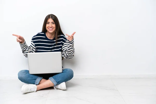 年轻的高加索女人 带着笔记本电脑坐在地板上 与白色背景隔离 手指指向侧边 快乐极了 — 图库照片