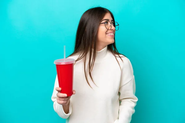 青い背景に隔離された若い白人女性飲みソーダ側を見て笑顔 — ストック写真