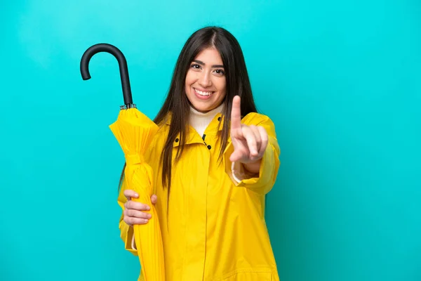 年轻的高加索女人 身穿防雨外套 带着雨伞 与蓝色背景隔离 露出并举起一个手指 — 图库照片