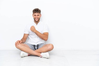 Genç sarışın adam, beyaz arka planda izole bir şekilde yerde oturmuş zaferi kutluyor.