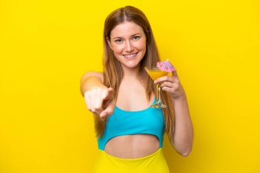 Sarı arka planda izole edilmiş bir kokteyl tutan genç beyaz kadın kendine güvenen bir ifadeyle sana işaret ediyor.