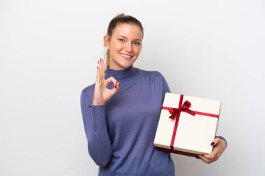 Beyaz arka planda izole edilmiş bir hediye tutan genç beyaz kadın parmaklarıyla tamam işareti gösteriyor.