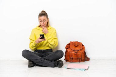 Beyaz arka planda beyaz bir zeminde oturan beyaz tenli genç bir kadın düşünüp bir mesaj gönderiyor.