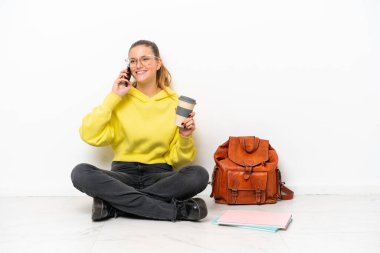 Beyaz arka planda bir katta oturan beyaz tenli genç bir kadın kahve alıp götürmek için elinde bir cep telefonu var.