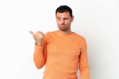 İzole edilmiş beyaz arka planı olan Brezilyalı adam mutsuz ve kenarı işaret ediyor.