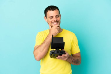 Brazilian man holding a drone remote control over isolated blue background looking to the side and smiling
