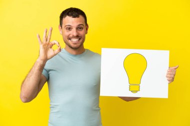 İzole edilmiş mor arkaplan üzerinde Brezilyalı bir adam elinde üzerinde ok işareti olan ampul simgesi olan bir levha tutuyor.