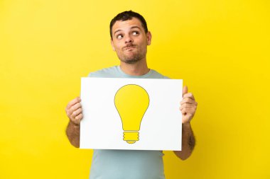 İzole edilmiş mor arka planı olan Brezilyalı bir adam elinde ampul simgesi olan bir levha tutuyor ve yukarı bakıyor.