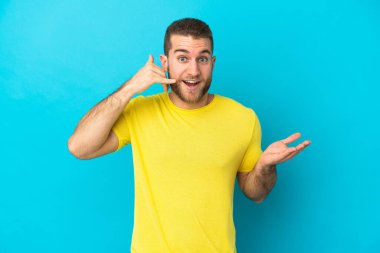 Genç yakışıklı beyaz adam mavi arka planda izole edilmiş telefon jestleri yapıyor ve şüpheli.