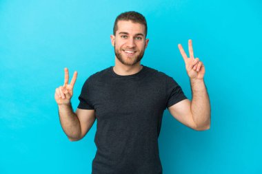 Mavi arka planda izole edilmiş yakışıklı beyaz adam iki eliyle zafer işareti gösteriyor.