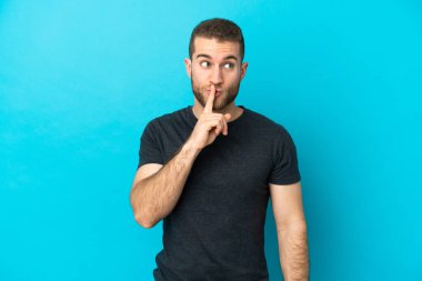 Mavi arka planda izole edilmiş yakışıklı beyaz adam sessiz bir jest işareti gösteriyor. Parmağını ağzına sokuyor.