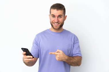 Young handsome caucasian man isolated on white background using mobile phone and pointing it