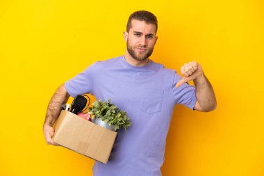 Genç Kafkasyalı, sarı arka planda izole edilmiş şeylerle dolu bir kutuyu seçerken negatif ifadeyle baş parmağını aşağı gösteriyor.