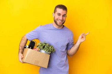 Genç Kafkasyalı, sarı arka planda izole edilmiş şeylerle dolu bir kutuyu kaldırırken hareket ediyor.