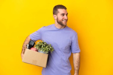 Genç Kafkasyalı, sarı arka planda izole edilmiş eşyalarla dolu bir kutuyu kaldırırken hamle yapıyor ve gülümsüyor.