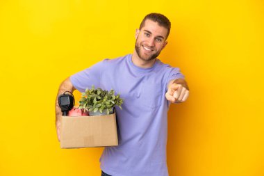 Genç Kafkasyalı, sarı arka planda izole edilmiş eşyalarla dolu bir kutuyu alıp mutlu bir ifadeyle öne doğru hamle yapıyor.