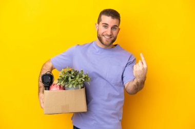 Genç Kafkasyalı, sarı arka planda izole edilmiş eşyalarla dolu bir kutuyu seçerken harekete geçiyor.