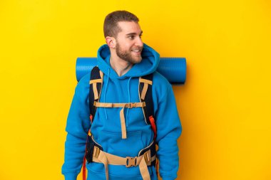 Sarı arka planda izole edilmiş büyük bir sırt çantası olan beyaz dağcı yan tarafa bakıyor ve gülümsüyor.