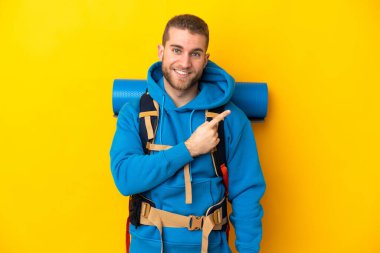 Sarı arka planda izole edilmiş büyük sırt çantalı beyaz bir dağcı bir ürünü sunmak için kenarı işaret ediyor.