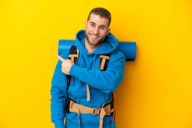 Young caucasian mountaineer man with a big backpack isolated on yellow background pointing back