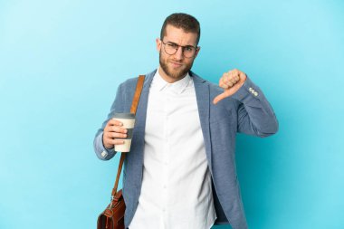 Kafkasyalı genç işadamı mavi arka planda izole edilmiş, olumsuz ifadeyle baş parmağını aşağı gösteriyor.