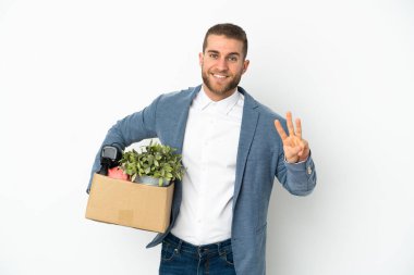 Beyaz arka planda izole edilmiş eşyalarla dolu bir kutuyu alırken harekete geçen beyaz tenli genç mutlu ve parmaklarıyla üçe kadar sayıyor.