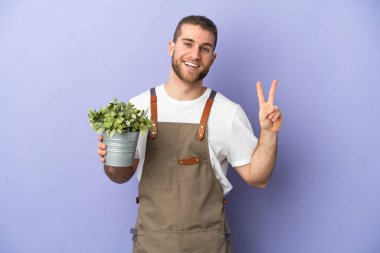 Bahçıvan beyaz adam sarı arka planda izole edilmiş bir bitki tutuyor gülümsüyor ve zafer işareti gösteriyor.