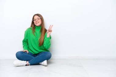 Beyaz arka planda izole bir şekilde yerde oturan genç beyaz kadın gülümsüyor ve zafer işareti gösteriyor.