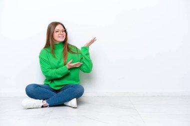 Beyaz arka planda izole bir şekilde yerde oturan beyaz tenli genç kadın davet edilmek için ellerini yana uzatıyor.