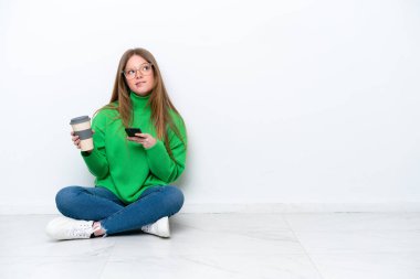 Beyaz arka planda izole bir şekilde yerde oturan beyaz tenli genç bir kadın elinde kahve ve bir şeyler düşünürken bir cep telefonu tutuyordu.