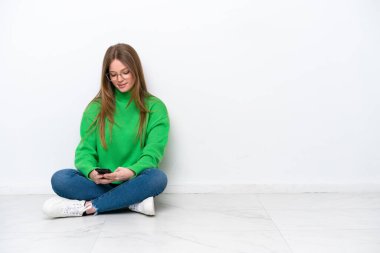 Beyaz arka planda izole bir şekilde yerde oturan beyaz bir kadın cep telefonuyla bir mesaj gönderiyor.