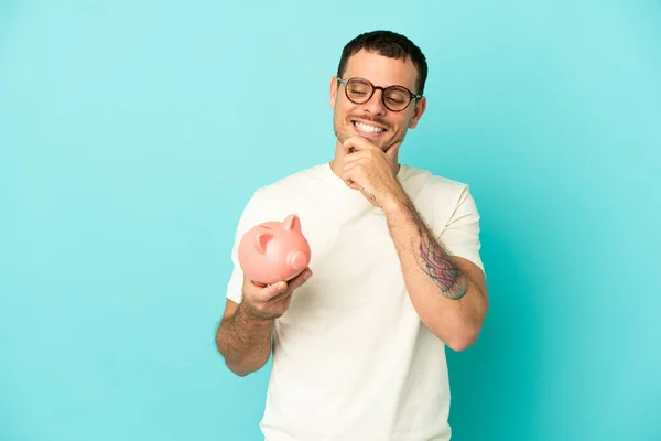 Brazilian Man Holding Piggybank Isolated Blue Background Looking Side Smiling — Stockfoto