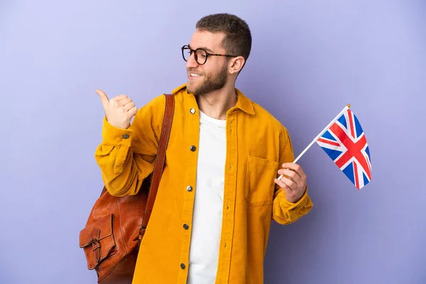 年轻的高加索人拿着一面英国国旗 用紫色背景隔开 指着侧面展示一种产品 — 图库照片