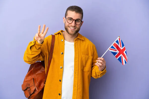 Young caucasian man holding an United Kingdom flag isolated on purple background happy and counting three with fingers