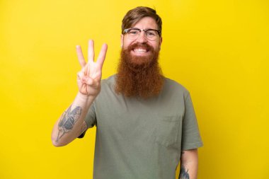 Sarı arka planda sakallı kızıl saçlı adam mutlu ve parmaklarıyla üçe kadar sayıyor.