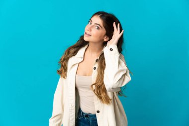 Mavi arka planda izole edilmiş genç beyaz kadın elini kulağına koyarak bir şeyler dinliyor.