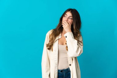 Mavi arka planda izole edilmiş genç beyaz kadın yan tarafa bakarken sürpriz jest yapıyor.