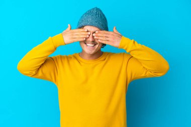 Yakışıklı sarışın adam mavi arka planda izole edilmiş gözlerini elleriyle kapatmış ve gülümsüyor.