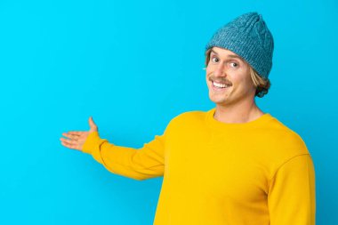 Yakışıklı sarışın adam mavi arka planda izole edilmiş. Davet etmek için ellerini yana uzatıyor.