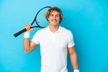 Genç sarışın adam mavi arka planda izole edilmiş tenis oynuyor.