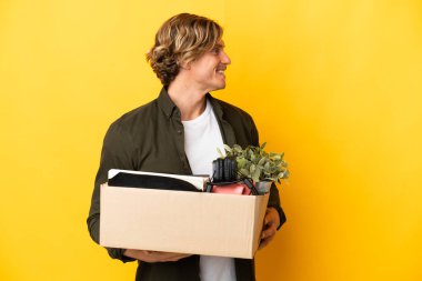 Sarışın adam, sarı arka planda izole edilmiş eşyalarla dolu bir kutuyu kaldırırken hamle yapıyor.