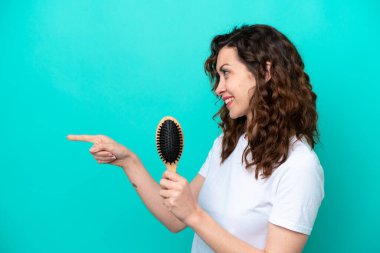Young caucasian woman holding hairbrush isolated on blue background pointing to the side to present a product