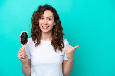 Mavi arka planda saç fırçası tutan beyaz bir kadın bir ürünü sunmak için yan tarafı işaret ediyor.