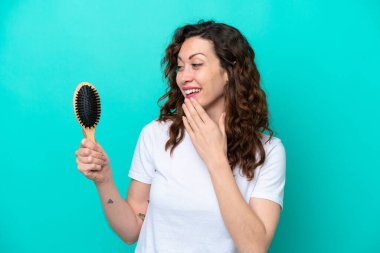 Mavi arka planda saç fırçası tutan beyaz bir kadın. Şaşırmış ve şok olmuş bir yüz ifadesi var.