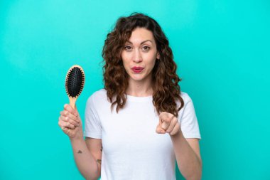 Mavi arka planda saç fırçası tutan genç beyaz kadın şaşırmış ve ön tarafı işaret ediyor.