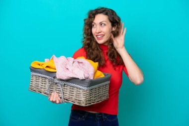 Kafkasyalı genç bir kadın mavi arka planda izole edilmiş bir çamaşır sepetini tutuyor ve elini kulağına koyup bir şeyler dinliyor.