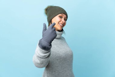 Kışlık şapkalı genç Uruguaylı kız mavi arka planda izole edilmiş. Gelmeye davet ediliyor.
