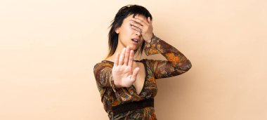 Hintli kadın bej arka planda izole edilmiş. Durma hareketi yapıyor ve yüzünü kapatıyor.