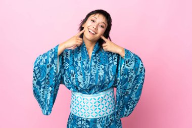 İzole edilmiş mavi arka planda kimono giyen genç bir kadın mutlu ve hoş bir ifadeyle gülümsüyor.