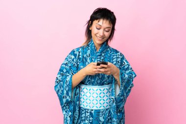 Kimono giyen genç bir kadın izole edilmiş mavi arka planda cep telefonuyla bir mesaj gönderiyor.